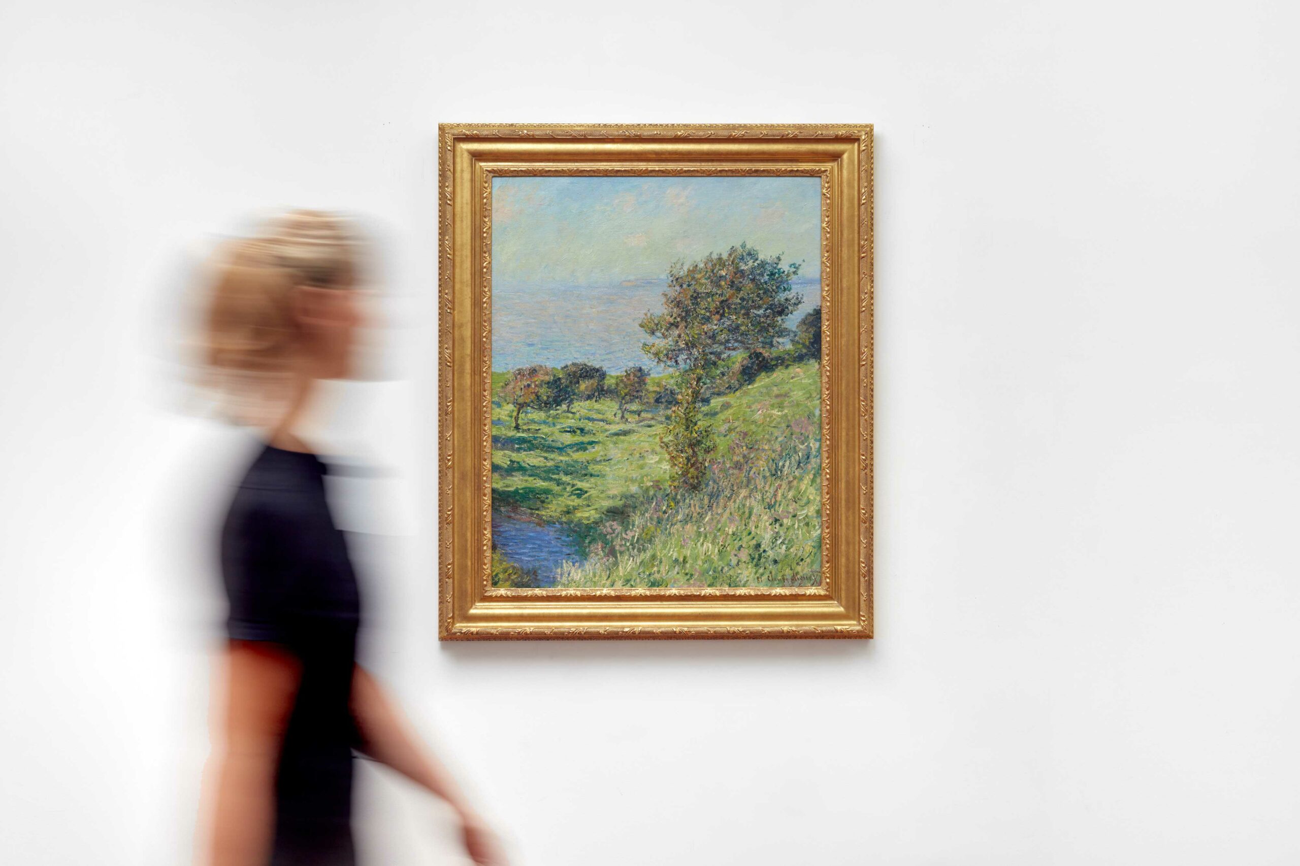 An impressionistic landscape painting by Monet on a white wall with a blurry viewer looking at it.