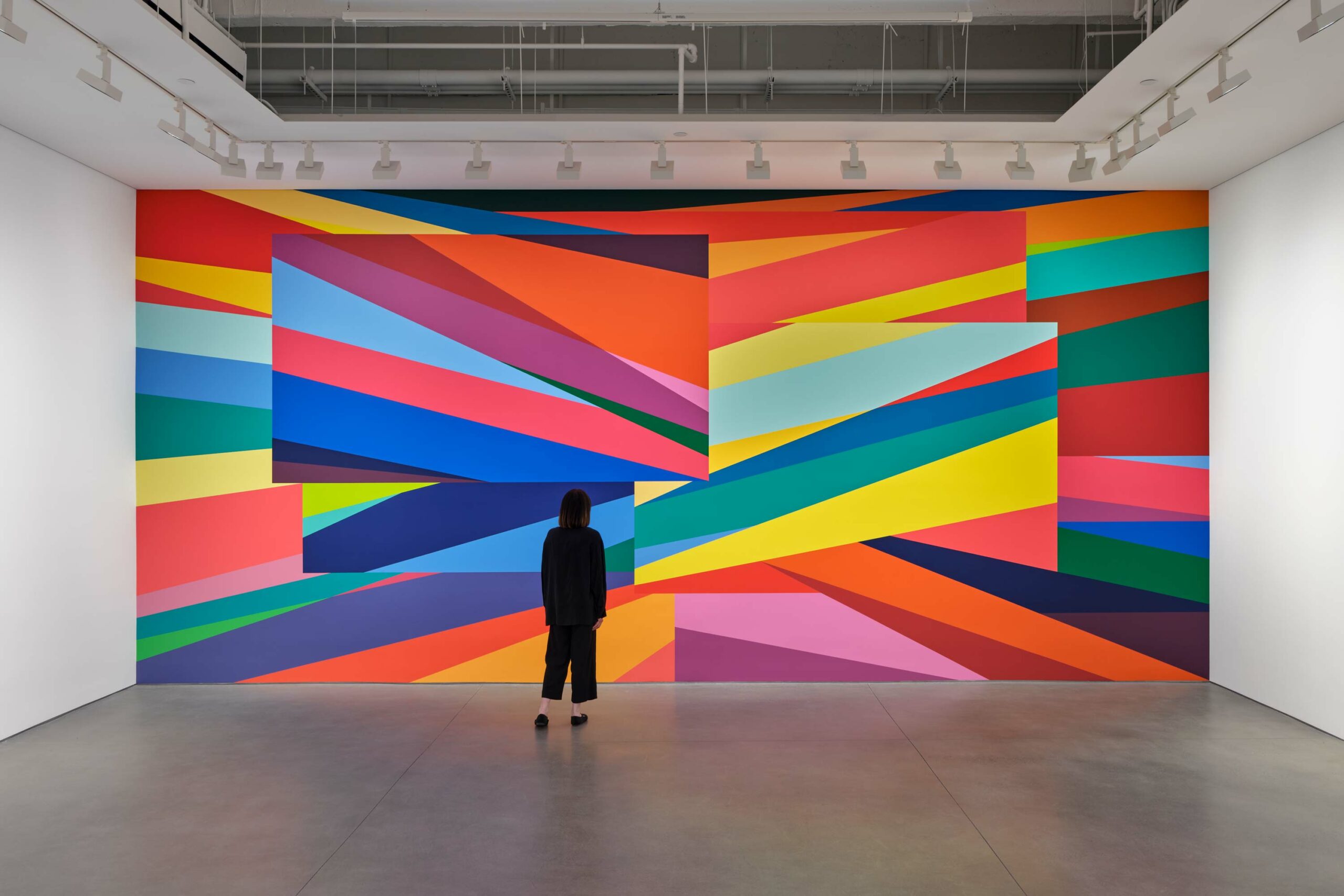 Large vibrant mural painting on a gallery. Abstract geometric blocks of color with a viewer looking at the artwork.