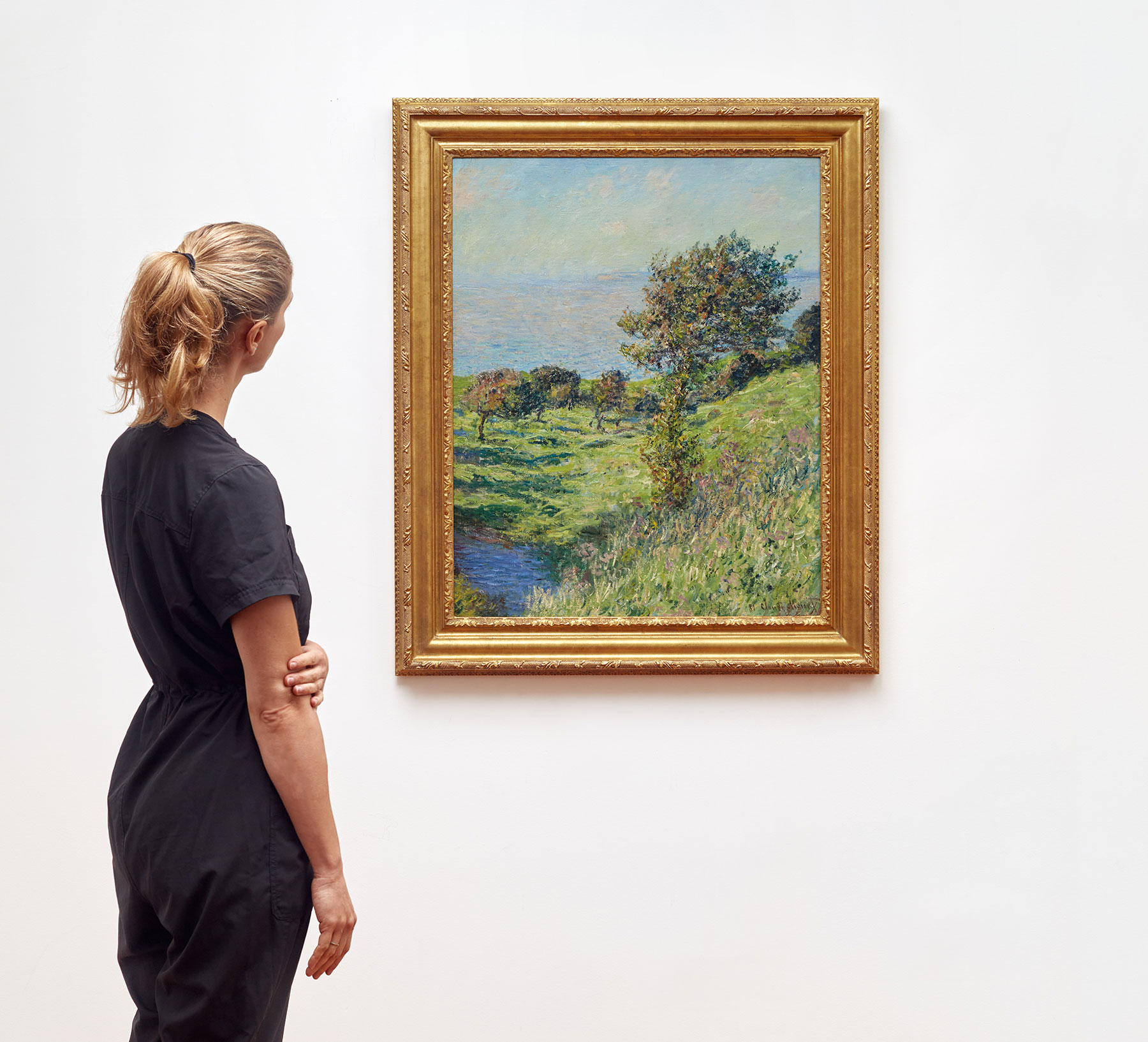 An impressionistic landscape painting by Monet in a white wall with a blonde viewer looking at it.