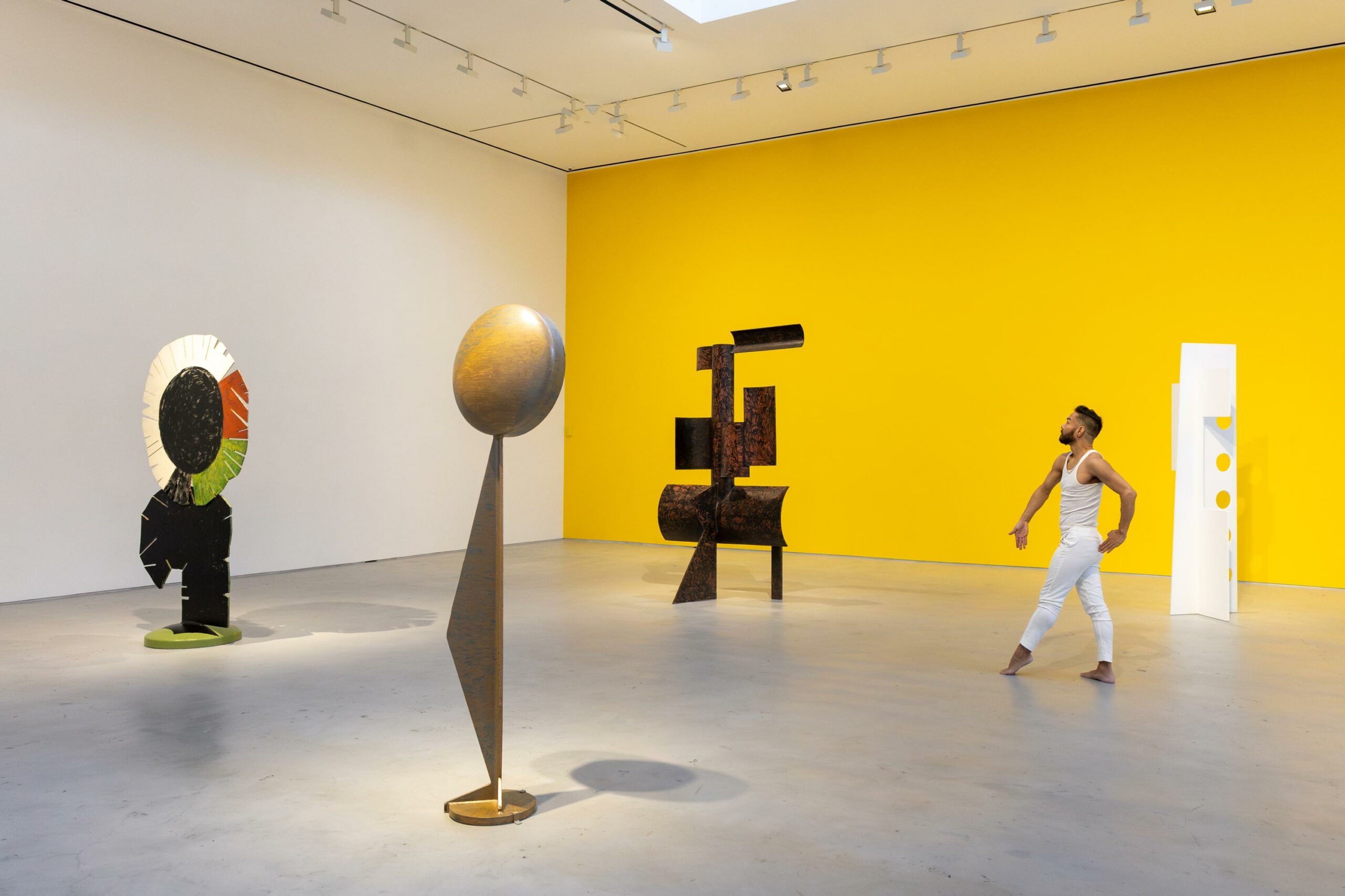 Wide exhibition view of 4 large geometric sculptures and a model wearing white posing still in front of a bright yellow wall.